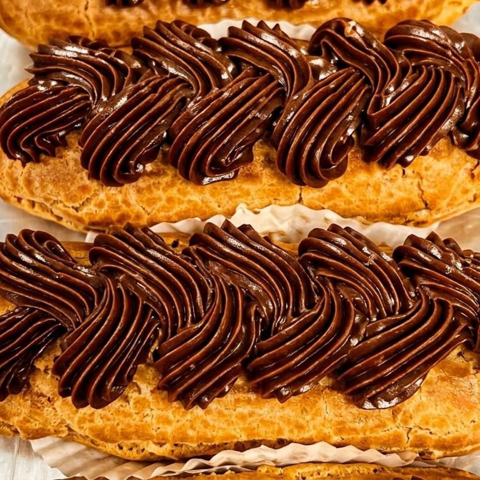 Rows of golden-brown eclairs topped with rich, piped chocolate frosting on a metal tray.